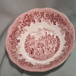 May include: A round ceramic bowl with a scalloped edge, featuring a pink and white transferware pattern. The center depicts a detailed scene of a historic building and a person on horseback, surrounded by a border of floral and berry motifs.