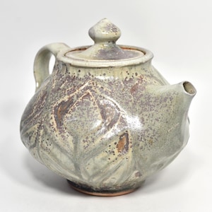 May include: A ceramic teapot with a rounded body and a matching lid. The surface features a mottled sage green and brown glaze with a textured, embossed leaf pattern across the exterior. It includes a curved handle and a short spout.