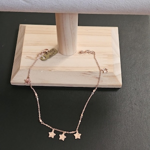 Puede incluir: Collar de color oro rosa con tres colgantes de estrella. El collar se exhibe en un expositor de joyas de madera. La cadena tiene pequeñas cuentas y es delicada.