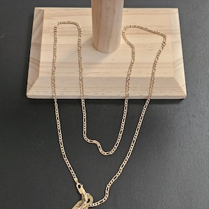 May include: A gold-colored Figaro chain necklace with a small tag pendant. The necklace is displayed on a wooden jewelry stand against a dark background. The tag pendant has engraved text.