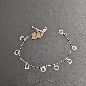 Könnte beinhalten: Ein filigranes Silberarmband mit einer Reihe kleiner, offener Kreise, die an einer feinen Kette hängen. Das Armband wird auf einer dunklen Oberfläche mit einem Preisschild gezeigt. Das Design des Armbands ist schlicht und elegant.