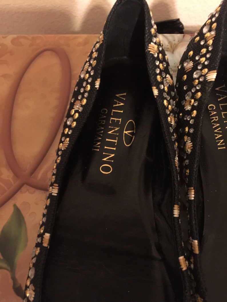May include: A pair of black flats with gold and silver embellishments. The shoes have the Valentino Garavani logo on the inside.