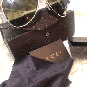 May include: White aviator sunglasses with brown lenses in a brown Gucci case with a brown Gucci cleaning cloth. The sunglasses have a gold trim and the case has a repeating Gucci logo pattern.
