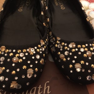May include: A pair of black ballet flats with gold and silver embellishments. The flats have the brand name "Valentino" written on the inside.
