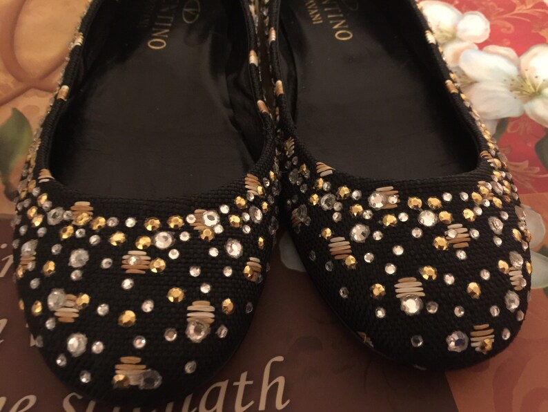 May include: A pair of black ballet flats with gold and silver embellishments. The flats have the brand name "Valentino" written on the inside.