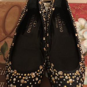 May include: A pair of black ballet flats with gold and silver embellishments. The shoes have the text "VALENTINO GARAVANI" on the inside.