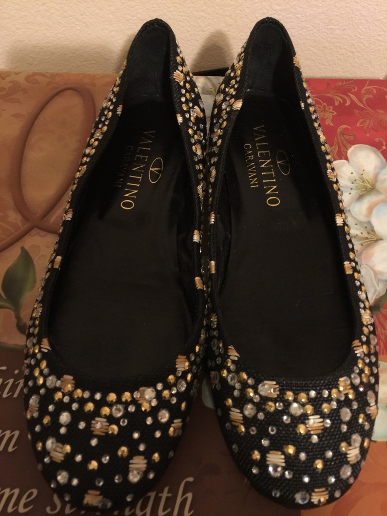 May include: A pair of black ballet flats with gold and silver embellishments. The shoes have the text "VALENTINO GARAVANI" on the inside.