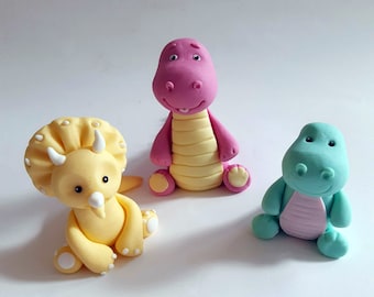 May include: Three small dinosaur figurines made of fondant. The dinosaurs are sitting on a white surface. The dinosaurs are yellow, pink, and green.
