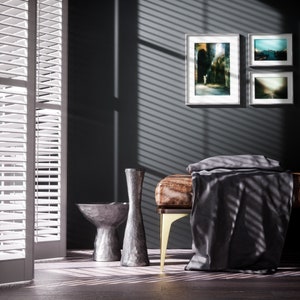 May include: A gray, leather bench with gold legs, a gray throw blanket, and two silver decorative vases. The bench is in front of a dark gray wall with three framed pictures.