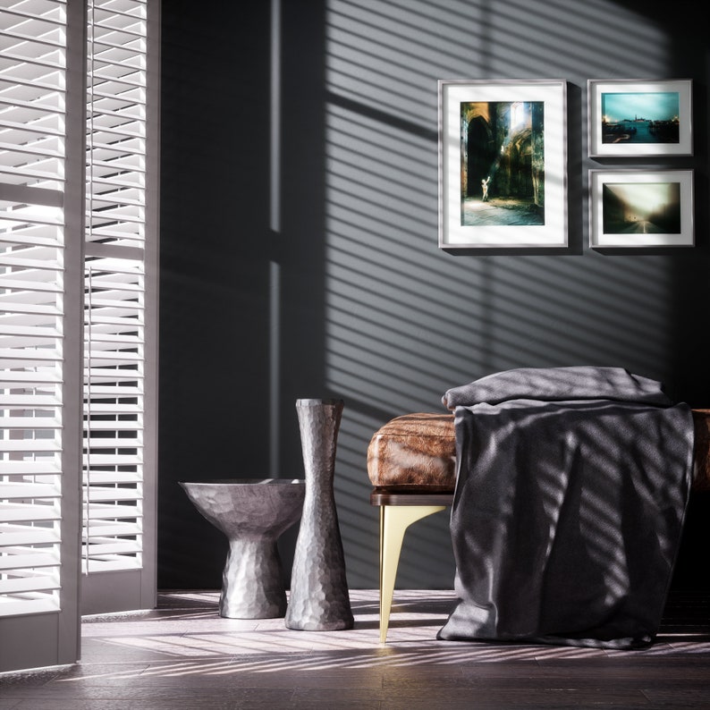 May include: A gray, leather bench with gold legs, a gray throw blanket, and two silver decorative vases. The bench is in front of a dark gray wall with three framed pictures.