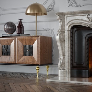Puede incluir: Un mueble de madera con paneles de puerta geom&eacute;tricos, coronado por una l&aacute;mpara dorada y objetos decorativos. El mueble tiene patas doradas. Una chimenea de m&aacute;rmol est&aacute; en el fondo y el suelo es de madera.