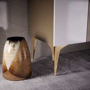 May include: A white and gold cabinet with gold legs. The cabinet is sitting on a gray carpet. A brown and white ceramic vase is in the foreground.