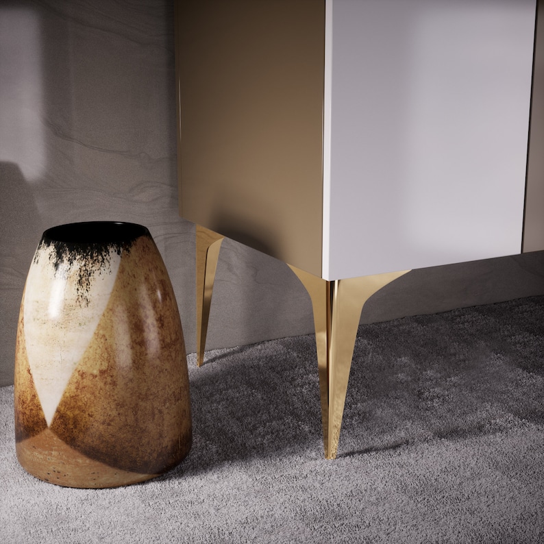 May include: A white and gold cabinet with gold legs. The cabinet is sitting on a gray carpet. A brown and white ceramic vase is in the foreground.