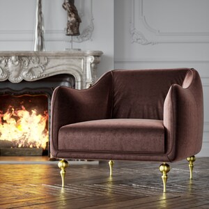 Puede incluir: Un sill&oacute;n de terciopelo marr&oacute;n con patas doradas. La silla tiene respaldo y reposabrazos redondeados. Las patas son ornamentadas y tienen un dise&ntilde;o bulboso. La silla est&aacute; frente a una chimenea con fuego.