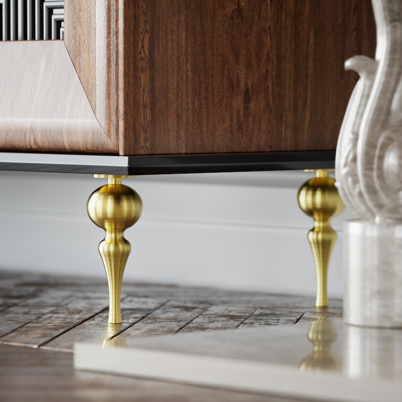 Puede incluir: Primer plano de un mueble de madera con patas de color lat&oacute;n. El mueble tiene un acabado de vetas de madera oscura y una moldura negra. Las patas son ornamentadas, con una parte superior en forma de bola y una base c&oacute;nica. El mueble est&aacute; sobre un suelo de madera.