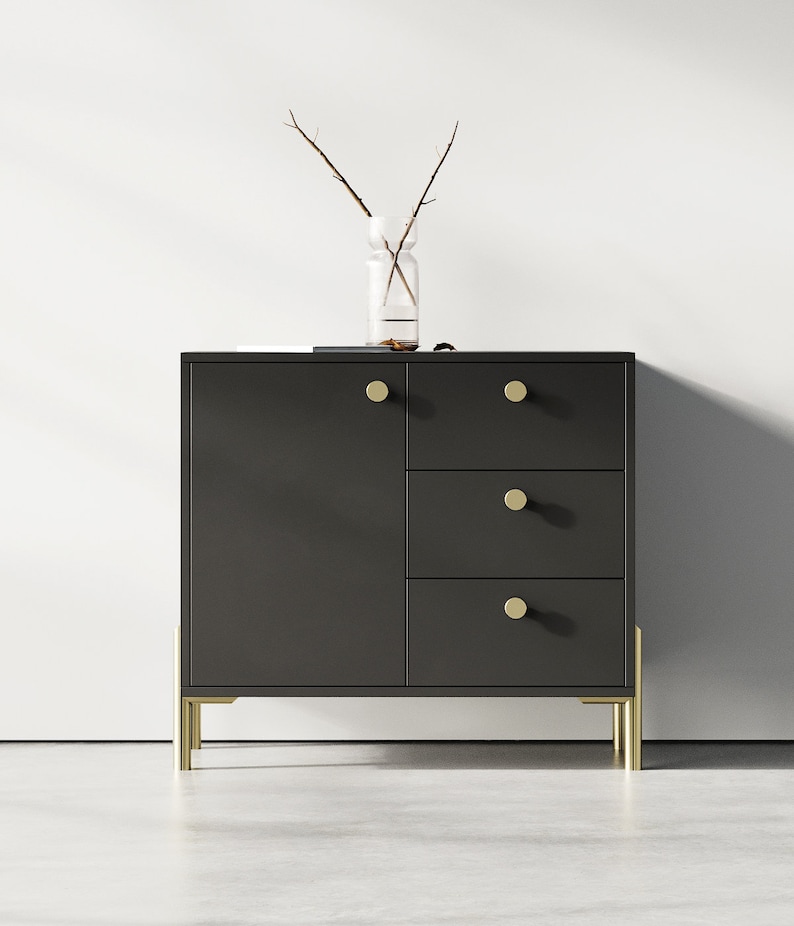 May include: A dark gray cabinet with three drawers and a door. The cabinet has gold metal legs and round gold knobs.