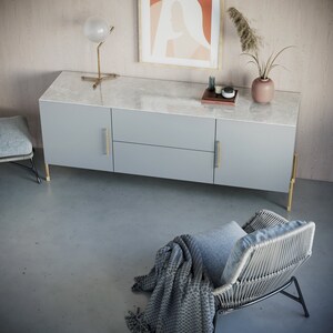 Puede incluir: Una c&oacute;moda gris claro con tres cajones y herrajes dorados. La c&oacute;moda est&aacute; sobre un suelo gris claro. Una peque&ntilde;a silla con un coj&iacute;n gris y una manta gris est&aacute; en primer plano.