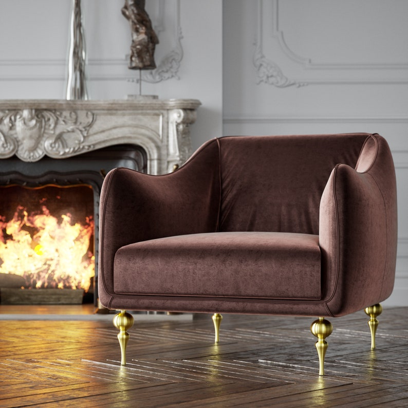 Puede incluir: Un sill&oacute;n de terciopelo marr&oacute;n con patas doradas. La silla tiene respaldo y reposabrazos redondeados. Las patas son ornamentadas y tienen un dise&ntilde;o bulboso. La silla est&aacute; frente a una chimenea con fuego.
