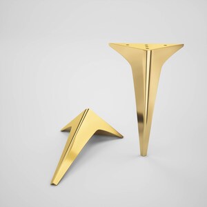 May include: Two gold metal furniture legs in a triangular shape. The legs are angled and have a pointed tip.