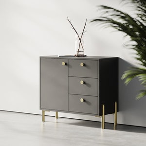 May include: A dark gray cabinet with three drawers and a door. The cabinet has gold metal legs and gold round knobs.