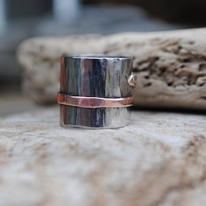 Könnte beinhalten: Ein silberner Ring mit gehämmerter Textur und einem Kupferband um die Mitte. Der Ring liegt auf einem Stück Treibholz.