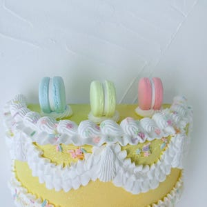 May include: A yellow cake decorated with white frosting, pastel sprinkles, and three macarons in blue, green, and pink. The cake has a ruffled frosting edge and is set against a white background.