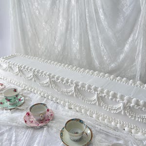 May include: A long, rectangular white cake decorated with intricate white piped frosting swags and borders. Three porcelain teacups with floral patterns and matching saucers are arranged in front of the cake on a white lace fabric surface.