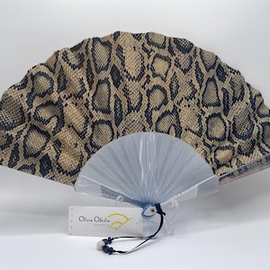 Peut inclure: Éventail décoratif avec un motif peau de serpent beige et noir. La base est transparente et plissée. Une étiquette indique "Olivia Oberlin". Un accessoire élégant pour se rafraîchir.