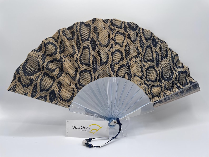 Pu&ograve; includere: Ventaglio decorativo con motivo a pelle di serpente beige e nero. Base trasparente e pieghettata. Etichetta con la scritta "Olivia Oberlin". Un accessorio elegante.