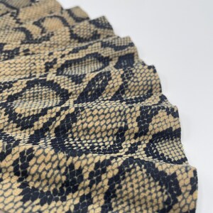 Pu&ograve; includere: Primo piano di un ventaglio con un motivo a pelle di serpente nei toni del beige e del nero. Il ventaglio &egrave; parzialmente aperto, rivelando il tessuto strutturato e il design intricato. Il logo Olivia Olivia &egrave; visibile sullo sfondo.