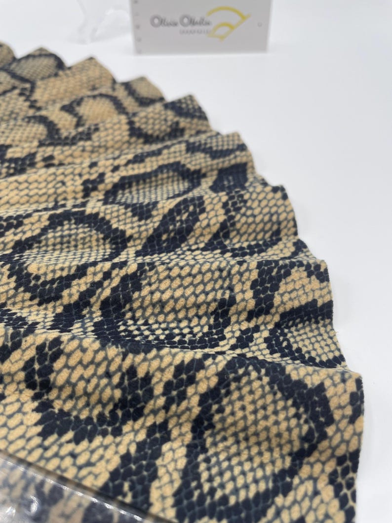 Pu&ograve; includere: Primo piano di un ventaglio con un motivo a pelle di serpente nei toni del beige e del nero. Il ventaglio &egrave; parzialmente aperto, rivelando il tessuto strutturato e il design intricato. Il logo Olivia Olivia &egrave; visibile sullo sfondo.