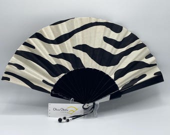 May include: A decorative hand fan with a black and white zebra stripe pattern. The fan has a black handle and a tag that reads "Olivia Oberlin essentials". The fan is open and displayed against a white background.