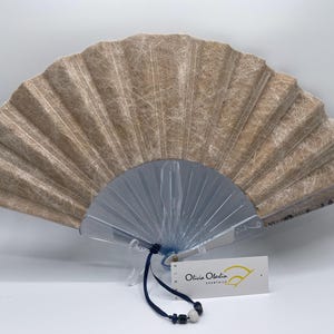 Pu&ograve; includere: Ventaglio di carta beige con base blu traslucido. Il ventaglio &egrave; aperto, mostrando il suo design a pieghe. Un'etichetta con il testo "Olivia Olerdin essentials" &egrave; appesa alla base. &Egrave; un oggetto decorativo.