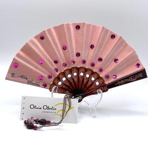 Peut inclure: Éventail rose avec un design décoratif. Le ventilateur a une texture plissée, ornée de paillettes violettes. La poignée est marron foncé avec un motif décoratif. Une étiquette avec le texte "Olivia Oberlin" est attachée. Une chaîne de perles est également présente.
