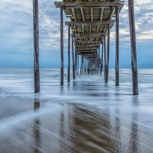 May include: A wooden pier extends out over a sandy beach with the ocean in the background. The water is a light blue and the sky is a cloudy gray. The pier is made of wooden planks and supports. The water is moving and creating a blurry effect.