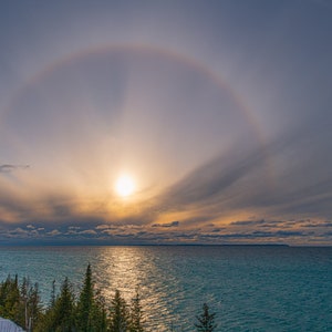 May include: A large halo, or ring of light, surrounds the sun in the sky above a body of water. The sun is shining brightly, and the water is a deep blue color. The sky is a light blue color, and there are some clouds in the sky.