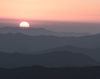 May include: A pink and orange sunset over a series of dark blue mountains.