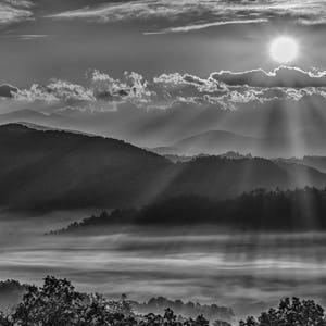 May include: Black and white landscape photograph of a mountain range at sunrise. The sun is visible, with rays of light shining through the clouds. The foreground features a misty valley and trees.