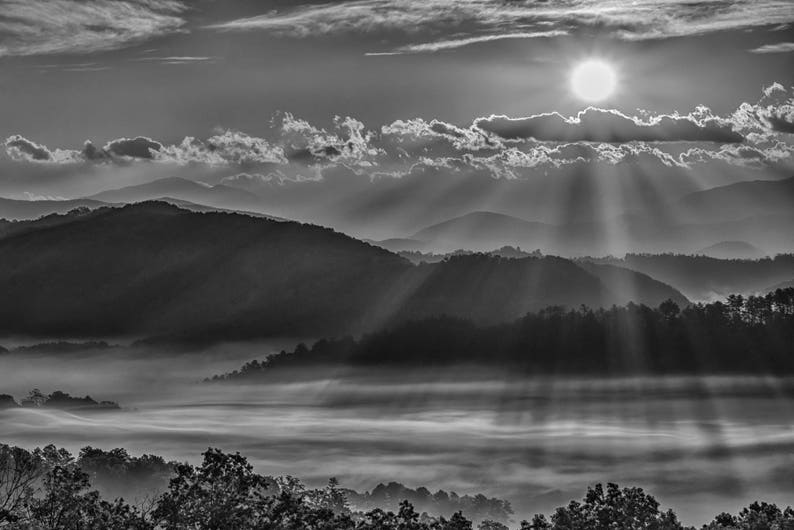 May include: Black and white landscape photograph of a mountain range at sunrise. The sun is visible, with rays of light shining through the clouds. The foreground features a misty valley and trees.