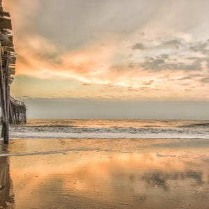 May include: A wooden pier extends out over a sandy beach with calm, clear water reflecting the cloudy sky. The sky is a mix of light blue, gray, and pink.