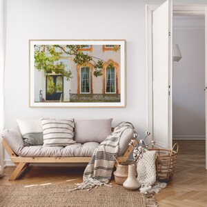 May include: A framed photograph of a building with a white facade and a green tree in front. The building has two windows with orange trim and a door with a black frame. The photograph is hanging on a white wall in a living room setting.