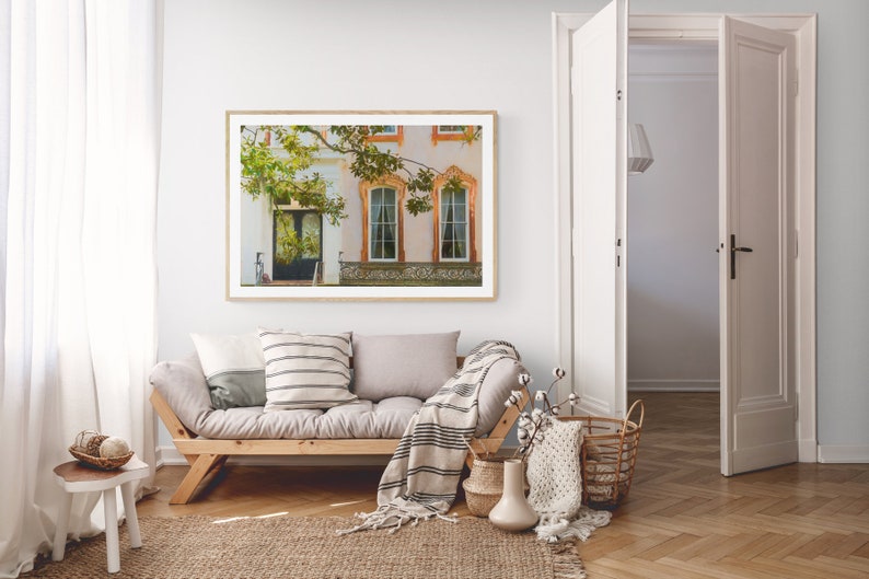 May include: A framed photograph of a building with a white facade and a green tree in front. The building has two windows with orange trim and a door with a black frame. The photograph is hanging on a white wall in a living room setting.
