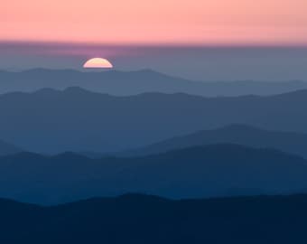 May include: A pink sunset over a series of blue mountains. The sun is setting behind the mountains, creating a soft, hazy glow.