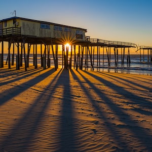 May include: A wooden pier with a building on top extends out over the ocean. The sun is setting behind the pier, casting long shadows on the sandy beach.