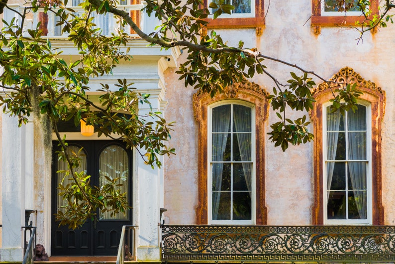 May include: A vintage-style building with a black double door and two large windows with white trim. The building has a balcony with an ornate iron railing. The building is painted in a light peach color and has a green tree in front of it.