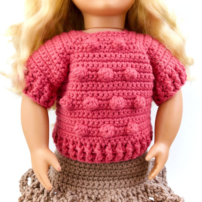 May include: A pink crocheted doll top with a textured bobble stitch pattern. The top has short sleeves and a round neckline.