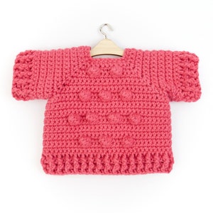 May include: A pink crocheted sweater with a textured design and short sleeves. The sweater is hanging on a wooden hanger.