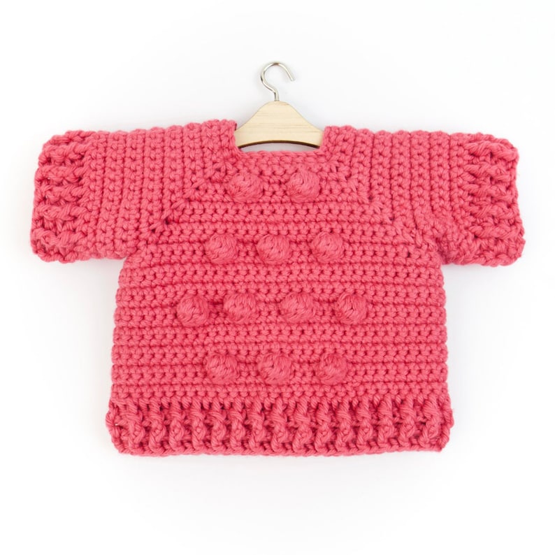 May include: A pink crocheted sweater with a textured design and short sleeves. The sweater is hanging on a wooden hanger.