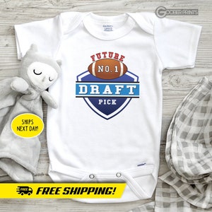 Puede incluir: Uniforme blanco para bebé con un gráfico de fútbol azul y rojo y el texto "FUTURE NO.1 DRAFT PICK".