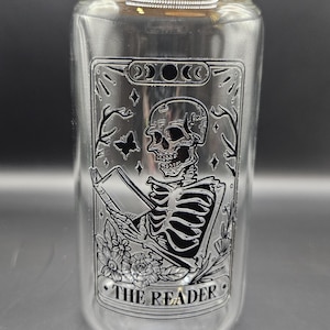 Puede incluir: Un vaso de vidrio transparente con tapa de bambú. El vaso presenta un gráfico negro de un esqueleto leyendo un libro, con las palabras "THE READER" en la parte inferior. El diseño incluye una mariposa, flores y fases lunares.